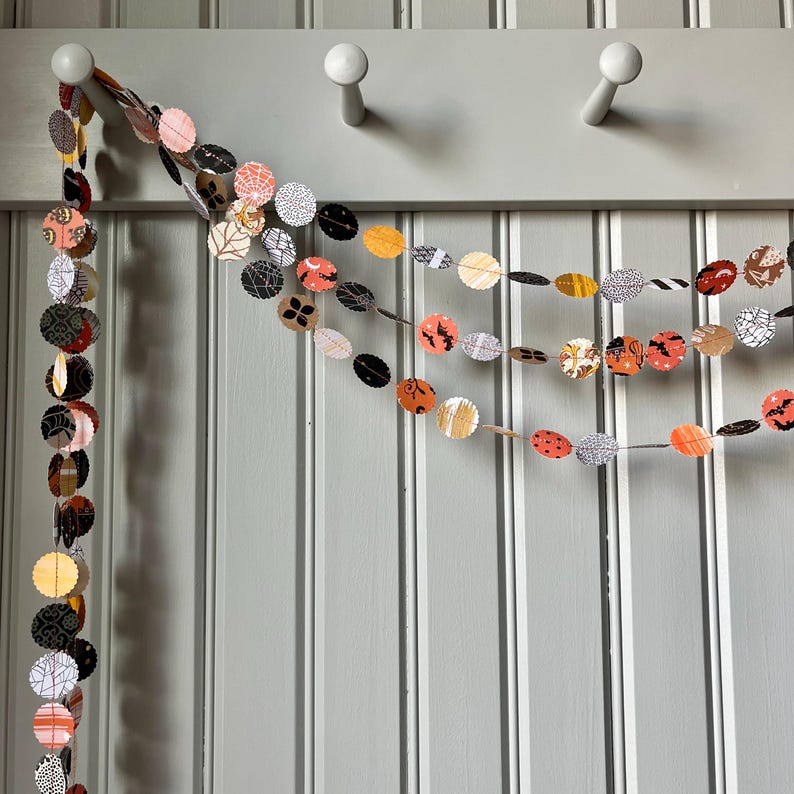 May include: A festive Halloween garland featuring circular paper cutouts in various patterns and colors, including orange, black, and white. The garland is strung together and hangs against a light gray wall, perfect for seasonal decor.