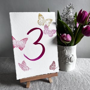 May include: A rectangular white card with a deckled edge featuring a large magenta number three in the center. The card is decorated with four butterfly illustrations in shades of gold and pink. It rests on a small wooden easel stand.