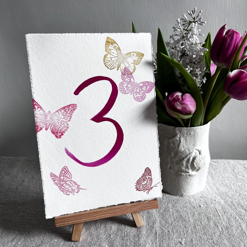 May include: A rectangular white card with a deckled edge featuring a large magenta number three in the center. The card is decorated with four butterfly illustrations in shades of gold and pink. It rests on a small wooden easel stand.