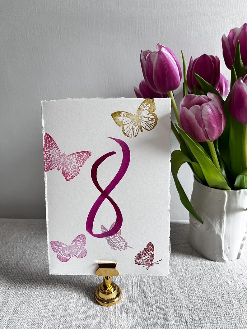 May include: A rectangular white card with deckled edges featuring a large magenta number 8 in the center, surrounded by several printed butterflies in shades of magenta and gold. The card is held upright by a small gold metal stand.