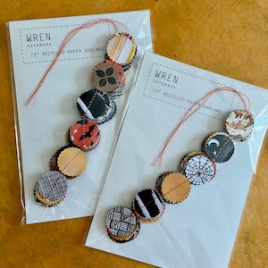 May include: Two packages of handmade paper garlands. Each garland is 72 inches long and features circular, patterned paper pieces in various colors and designs, including bats, spiderwebs, and geometric patterns. The text "wren handmade" is printed on the packaging.