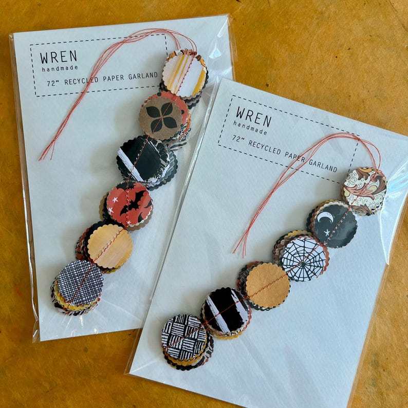 May include: Two packages of handmade paper garlands. Each garland is 72 inches long and features circular, patterned paper pieces in various colors and designs, including bats, spiderwebs, and geometric patterns. The text "wren handmade" is printed on the packaging.