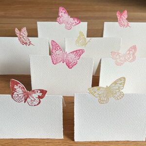 May include: A collection of white folded cards, each adorned with a different butterfly design. The butterflies are in shades of pink, red, and yellow. The cards are arranged on a wooden surface.