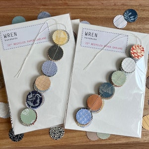 May include: Two white cards with the text "WREN handmade 72" RECYCLED PAPER GARLAND" on them. Each card has a string of colorful paper circles attached to it. The circles are made of different patterned papers and are strung together with white thread.