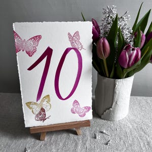 May include: A rectangular white card with deckled edges featuring the number 10 in a large magenta font. The card is decorated with four butterfly illustrations in shades of magenta and gold, and it is displayed on a small wooden easel.