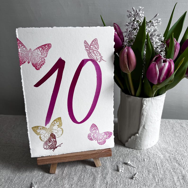 May include: A rectangular white card with deckled edges featuring the number 10 in a large magenta font. The card is decorated with four butterfly illustrations in shades of magenta and gold, and it is displayed on a small wooden easel.