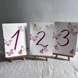 May include: Three rectangular white paper cards with deckled edges, each displaying a large dark pink number and several small butterflies in shades of pink, magenta, and gold. Each card is supported by a small wooden tripod easel stand.