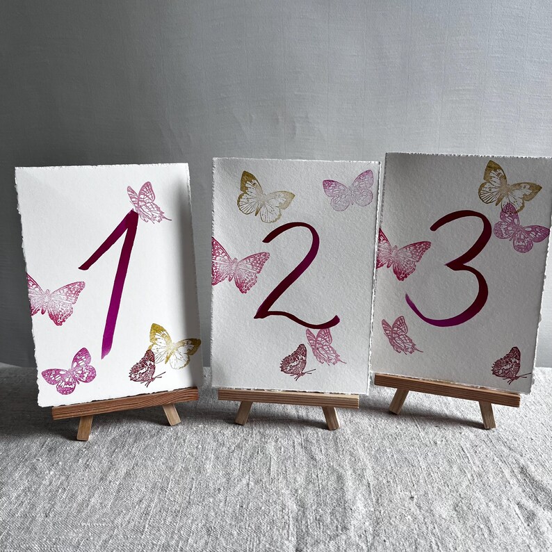 May include: Three rectangular white paper cards with deckled edges, each displaying a large dark pink number and several small butterflies in shades of pink, magenta, and gold. Each card is supported by a small wooden tripod easel stand.