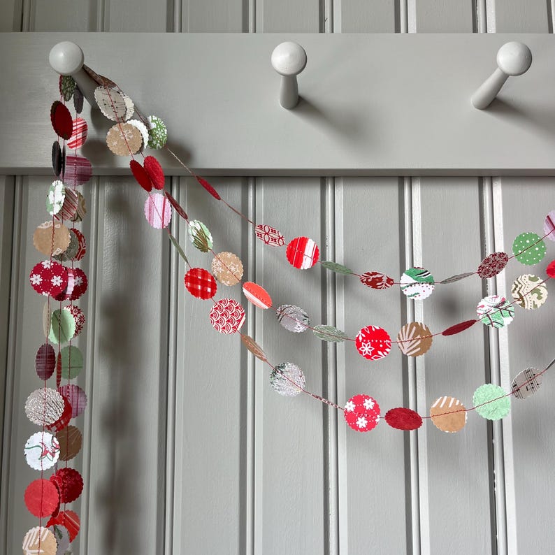 May include: A festive garland made of red, green, and brown paper circles with various patterns. The garland is strung on a white cord and is hanging from a white hook.