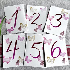 May include: Six rectangular white paper cards with deckled edges, each featuring a large dark magenta handwritten number from one to six. The cards are decorated with small, detailed butterfly illustrations in shades of magenta, pink, and gold.
