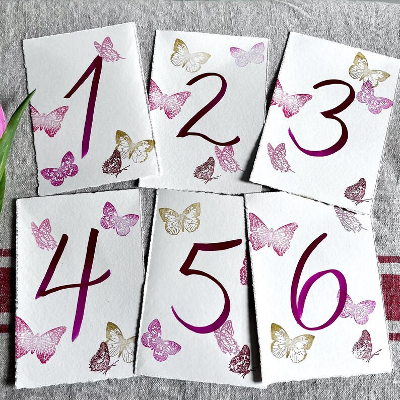May include: Six rectangular white paper cards with deckled edges, each featuring a large dark magenta handwritten number from one to six. The cards are decorated with small, detailed butterfly illustrations in shades of magenta, pink, and gold.