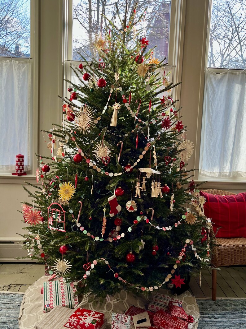 May include: A Christmas tree decorated with red ornaments, candy canes, and straw stars. The tree is in a room with a window and a wicker chair with a red pillow.