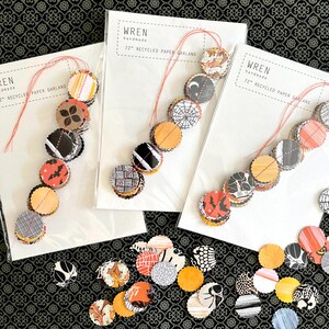 May include: Three handmade paper garlands with circular cutouts in various patterns and colors, including orange, black, and white. Each garland is 72 inches long and strung on red thread. The packaging reads "Wren handmade 72" Recycled Paper Garland."