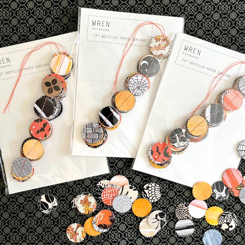 May include: Three handmade paper garlands with circular cutouts in various patterns and colors, including orange, black, and white. Each garland is 72 inches long and strung on red thread. The packaging reads "Wren handmade 72" Recycled Paper Garland."