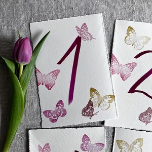 May include: White rectangular cards with deckled edges featuring the numbers 1 and 2 printed in a dark magenta ink. The cards are decorated with various butterfly illustrations in shades of magenta, pink, and gold. A single purple tulip is visible.
