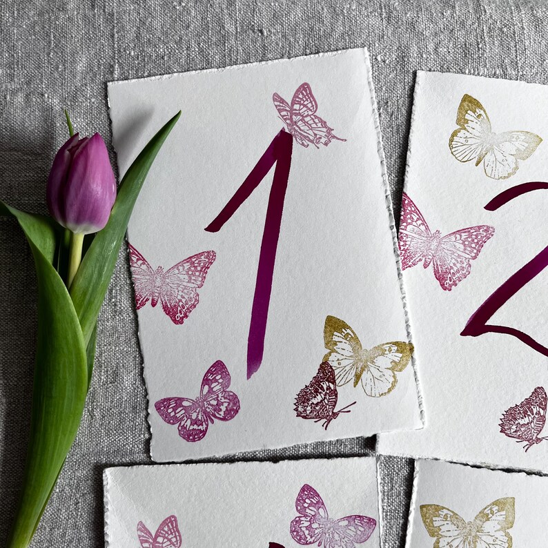 May include: White rectangular cards with deckled edges featuring the numbers 1 and 2 printed in a dark magenta ink. The cards are decorated with various butterfly illustrations in shades of magenta, pink, and gold. A single purple tulip is visible.