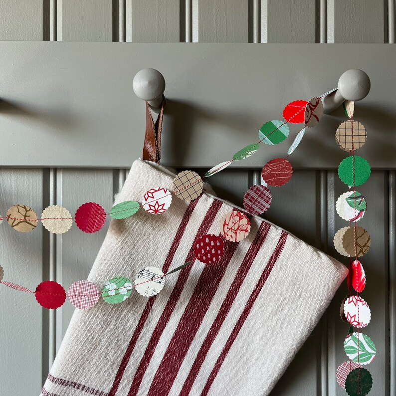 May include: A white and red striped stocking with a colorful fabric garland hanging from it. The garland is made of various shapes and patterns, including circles, squares, and stars. The stocking is hung on a hook on a gray wall.