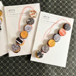 May include: Two handmade paper garlands, each 72 inches long, with circular paper cutouts in various patterns and colors. The text "WREN handmade" is printed on the white card. The garland features orange, black, and white designs, perfect for festive decor.
