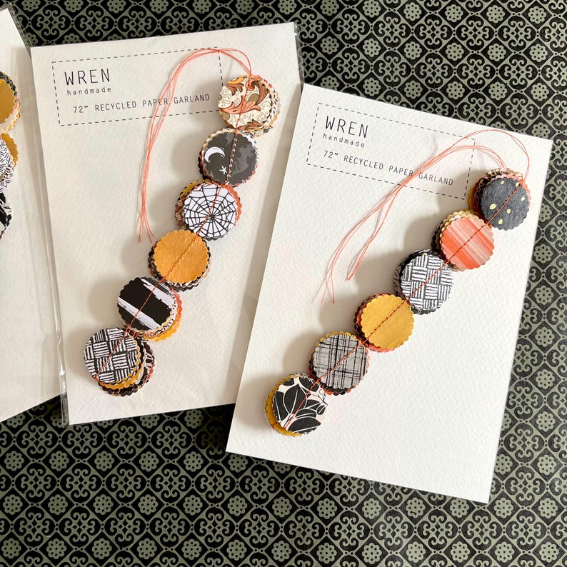 May include: Two handmade paper garlands, each 72 inches long, with circular paper cutouts in various patterns and colors. The text "WREN handmade" is printed on the white card. The garland features orange, black, and white designs, perfect for festive decor.