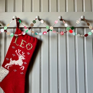 May include: A red knitted stocking with a white reindeer and the word "LED" on it hangs from a white hook. A paper garland with red, green, and white circles hangs above the stocking.