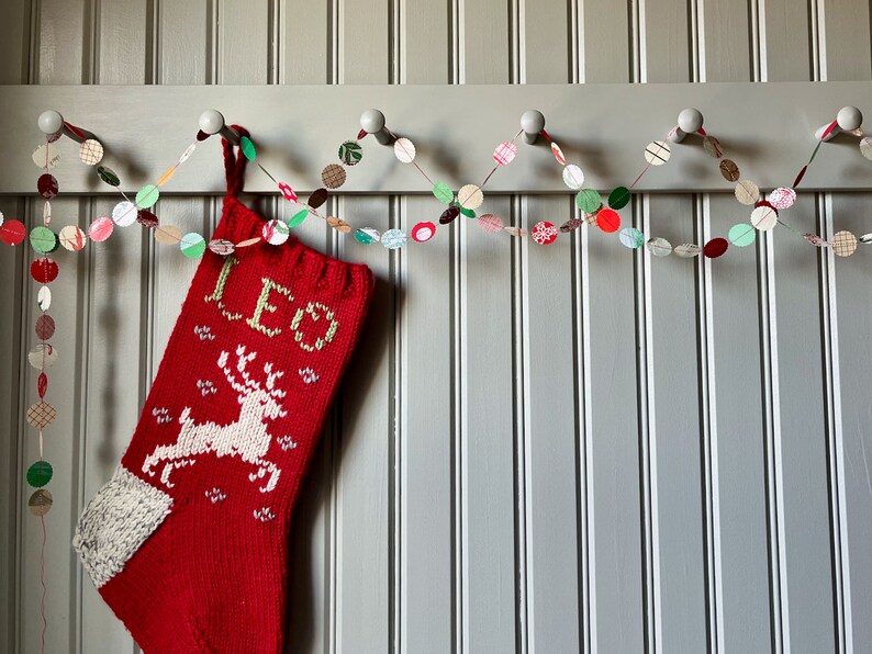 May include: A red knitted stocking with a white reindeer and the word "LED" on it hangs from a white hook. A paper garland with red, green, and white circles hangs above the stocking.
