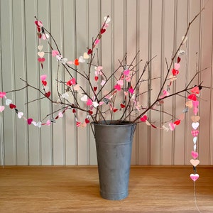 May include: A vase filled with branches decorated with paper hearts in shades of pink, red, and white. The vase is gray and sits on a wooden surface.