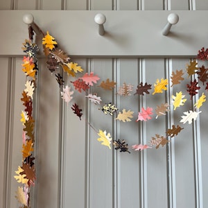 May include: A fall-themed leaf garland in shades of yellow, brown, pink, and white hangs against a gray wall. The garland features various leaf shapes and patterns, creating a festive and decorative display. The garland is strung on a thin brown string.