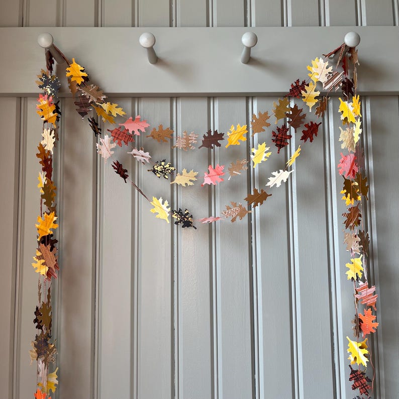 K&ouml;nnte beinhalten: Herbstliche Girlande mit Bl&auml;ttern aus Papier in verschiedenen Farben wie Gelb, Orange, Braun und Rosa. Die Bl&auml;tter sind an einer Schnur befestigt und h&auml;ngen an einer Wand mit wei&szlig;en Kn&ouml;pfen, ideal f&uuml;r Dekorationen.