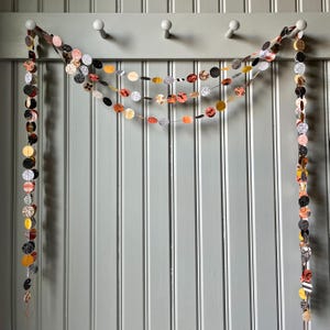 May include: A decorative garland featuring circular paper cutouts in various patterns and colors, including black, orange, white, and patterned designs. The garland is suspended from a row of white knobs against a gray wall, creating a festive and visually appealing display.