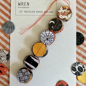 May include: A 72-inch recycled paper garland with various Halloween-themed designs, including a spiderweb, bats, and pumpkins. The garland is handmade and features a variety of colors and patterns. The card reads "wren handmade".
