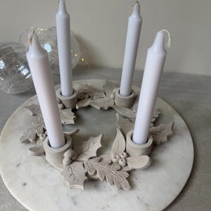 Puede incluir: Un portavelas circular de arcilla gris claro con cuatro soportes cilíndricos integrados para velas cónicas. La base está decorada con hojas de acebo y bayas esculpidas. Cuatro velas blancas están insertadas en los soportes.