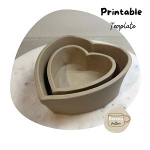 May include: Two heart-shaped ceramic bowls, one nested inside the other, on a white marble surface. The bowls are a light brown colour and have a smooth, slightly textured finish. The image includes the text "Printable Template" and "Pottery pattern".