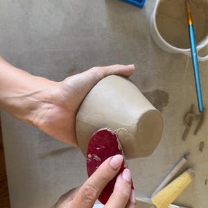 May include: A beige clay bowl being smoothed with a red rubber rib tool. Nearby are a white ceramic bowl containing slip, a blue plastic trimming tool, a yellow sponge on a wooden handle, and a small metal sculpting tool on a flat surface.