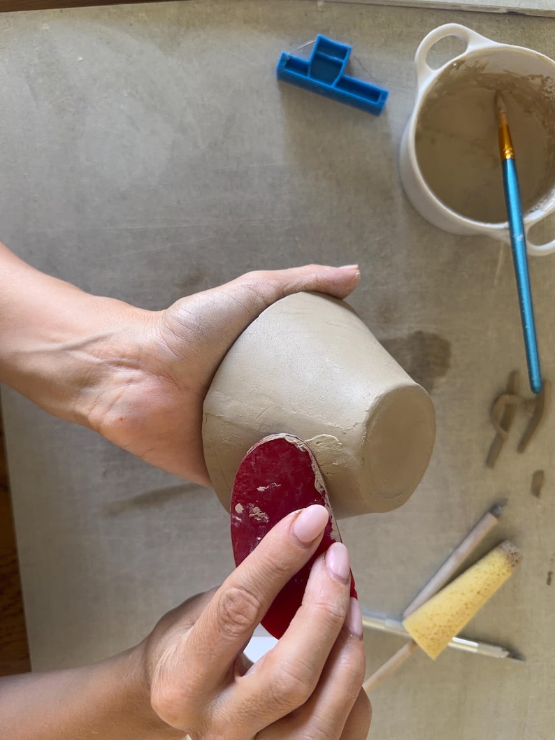 May include: A beige clay bowl being smoothed with a red rubber rib tool. Nearby are a white ceramic bowl containing slip, a blue plastic trimming tool, a yellow sponge on a wooden handle, and a small metal sculpting tool on a flat surface.