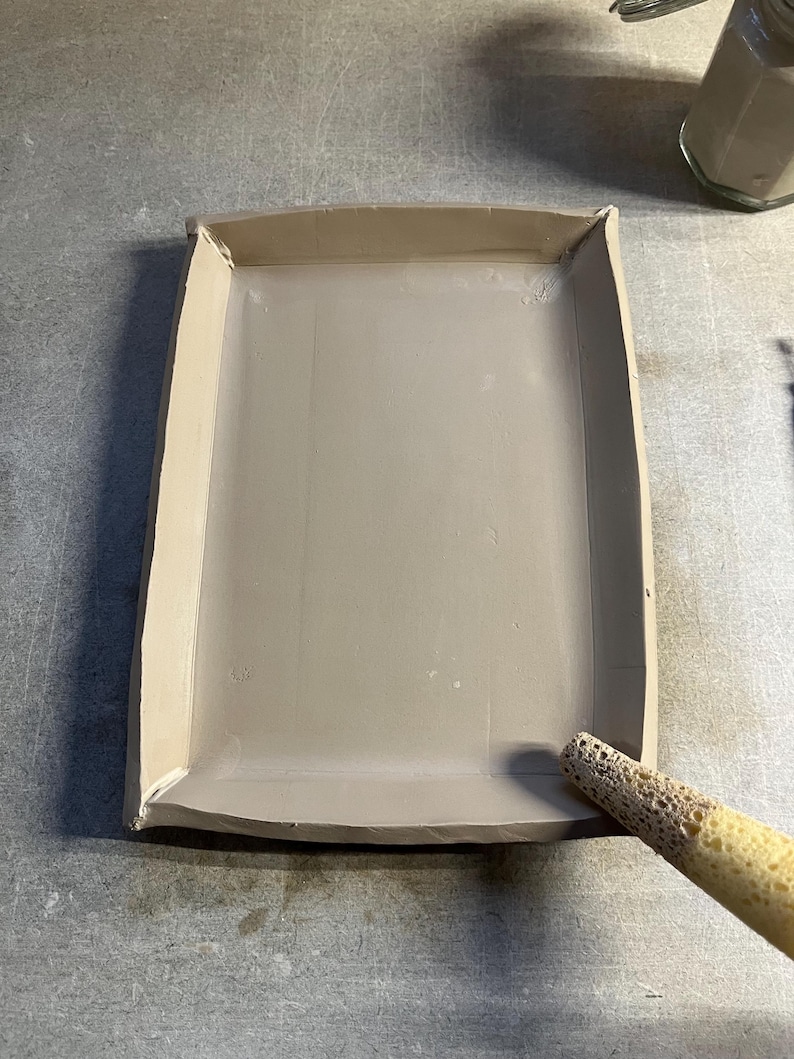K&ouml;nnte beinhalten: Ein hellbraunes Keramiktablett mit abgerundeten Kanten. Ein Schwammwerkzeug wird verwendet, um die Kanten des Tabletts zu gl&auml;tten.
