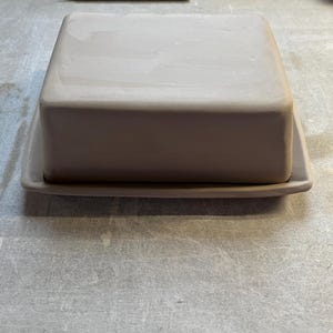 K&ouml;nnte beinhalten: Eine Butterschale aus Keramik mit Deckel und rechteckigem Boden. Die Schale ist hellbraun und hat eine glatte, matte Oberfl&auml;che.