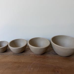 May include: A set of four smooth, matte, light beige ceramic bowls arranged in a row from smallest to largest. Each bowl features a simple, rounded shape with a consistent neutral color and a clean, unadorned surface throughout the entire set.