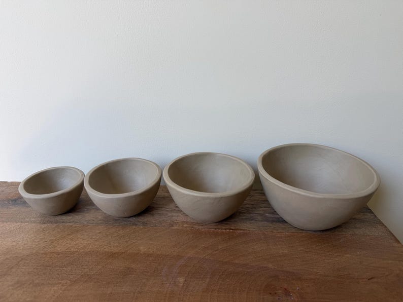 May include: A set of four smooth, matte, light beige ceramic bowls arranged in a row from smallest to largest. Each bowl features a simple, rounded shape with a consistent neutral color and a clean, unadorned surface throughout the entire set.