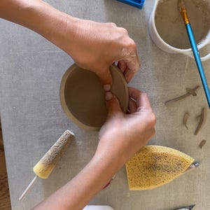 May include: A brown clay bowl being shaped, accompanied by a white ceramic container with slip, a blue wire clay cutter, a blue-handled paintbrush, a yellow sponge on a stick, a triangular yellow sponge, and a metal craft knife on a gray surface.
