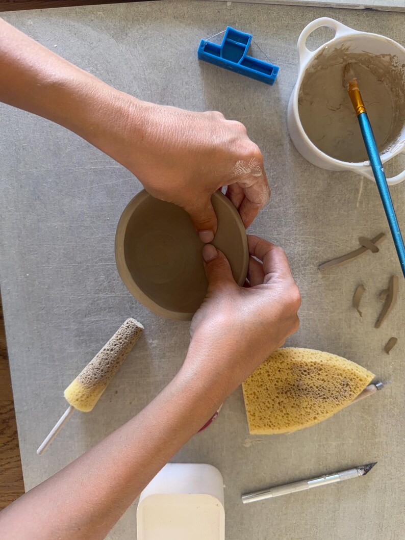 May include: A brown clay bowl being shaped, accompanied by a white ceramic container with slip, a blue wire clay cutter, a blue-handled paintbrush, a yellow sponge on a stick, a triangular yellow sponge, and a metal craft knife on a gray surface.