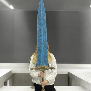 May include: A large, blue, decorative sword with intricate carvings and a gold-coloured hilt. The sword is held by a person in front of a white wall.