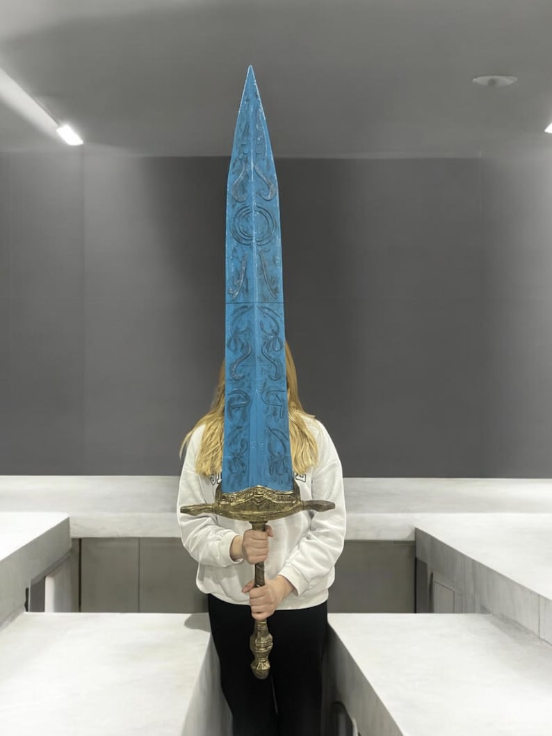 May include: A large, blue, decorative sword with intricate carvings and a gold-coloured hilt. The sword is held by a person in front of a white wall.