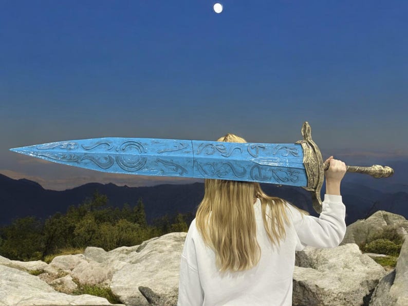 May include: A person is holding a large, blue, decorative sword with gold accents above their head. The sword has intricate designs carved into the blade. The person is standing on a rocky mountaintop with a view of a forest and mountains in the distance.