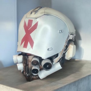 May include: A weathered, off-white helmet with a prominent red cross symbol on the front. The helmet features multiple cylindrical components and a detailed, industrial design, suggesting a futuristic or sci-fi theme.