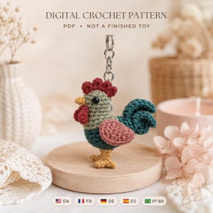 Op de afbeelding: Een digitaal haakpatroon voor een haan-sleutelhanger. De amigurumi-haan heeft een saliegroen lichaam, een donkerrode kam en lellen, een gele snavel en poten, een oudroze vleugel en een donkerblauwe staart, bevestigd aan een zilverkleurige metalen ring.