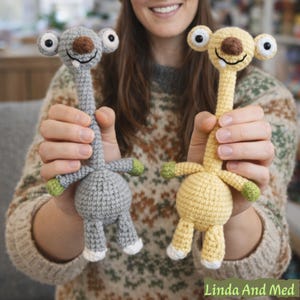 May include: Two handmade crochet toys, one gray and one yellow, are held up. Each has a long neck, large eyes, and a smiling face. The toys have small arms and legs with green and white accents. The toys are whimsical and soft.