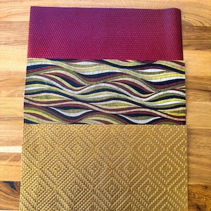 Op de afbeelding: Drie placemats met verschillende patronen. De bovenste placemat is effen bordeauxrood. De middelste placemat heeft een golvend patroon met gouden, bruine en zwarte strepen. De onderste placemat is geweven goudkleurig.