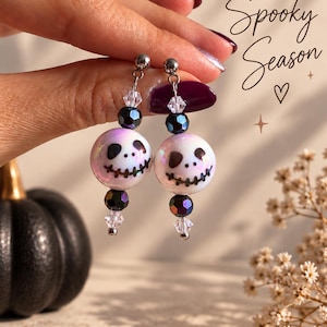Könnte beinhalten: Hängeohrringe mit irisierenden weißen Perlen, die mit schwarzen Totenkopfgesichtern bemalt sind, akzentuiert durch kleinere dunkelmetallische Perlen und klare facettierte Kristalle. Der Text Spooky Season ist sichtbar.
