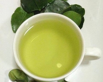 Puede incluir: Una taza de cerámica blanca llena de té verde claro, rodeada de hojas verdes de lima kaffir sobre una superficie blanca.