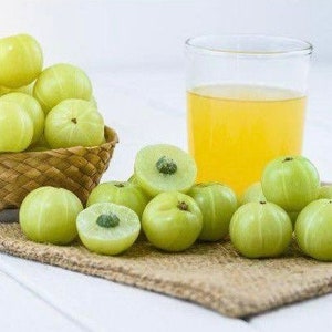 May include: A glass of yellow juice with green Indian gooseberries, also known as amla, on a brown hessian surface.